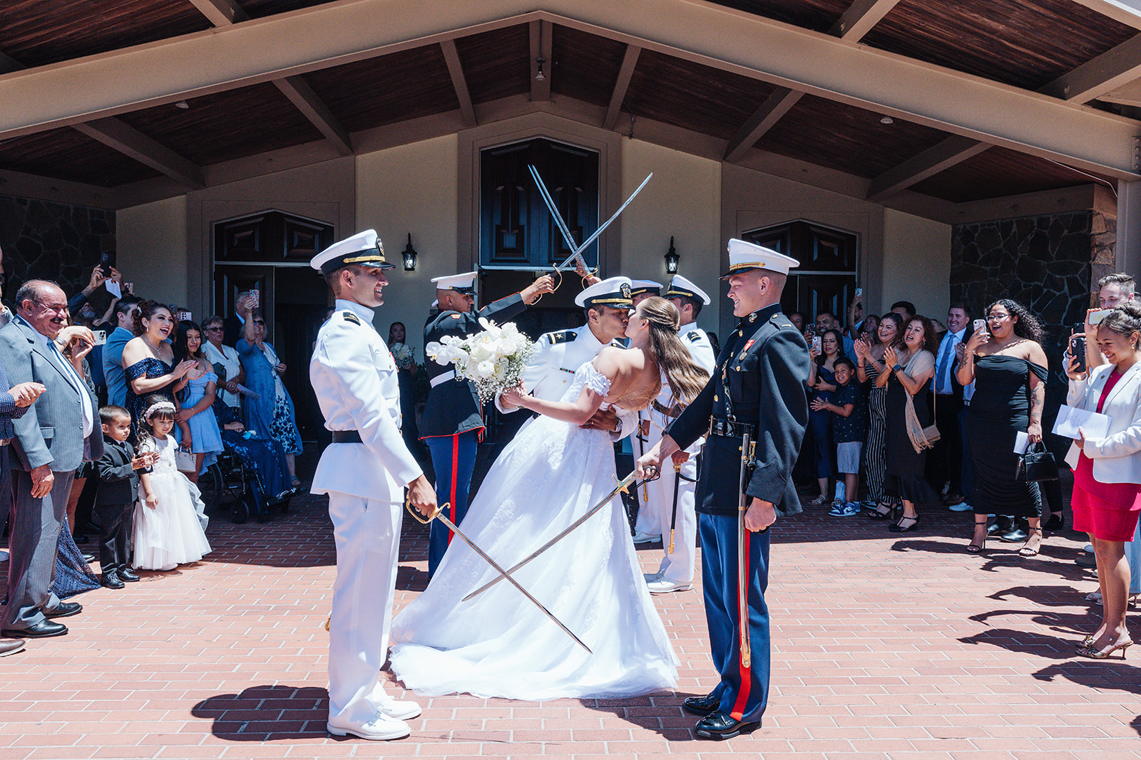Bride and groom share a kiss during a military saber arch exit as guests cheer outside a wedding ceremony