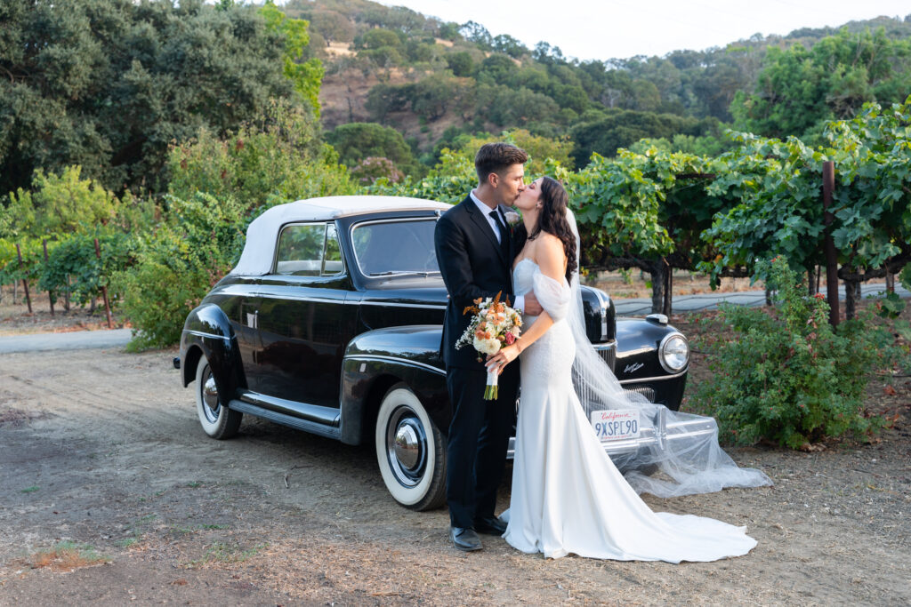 Bride and groom sharing a romantic moment next to a classic car in a vineyard during their wedding in California.
